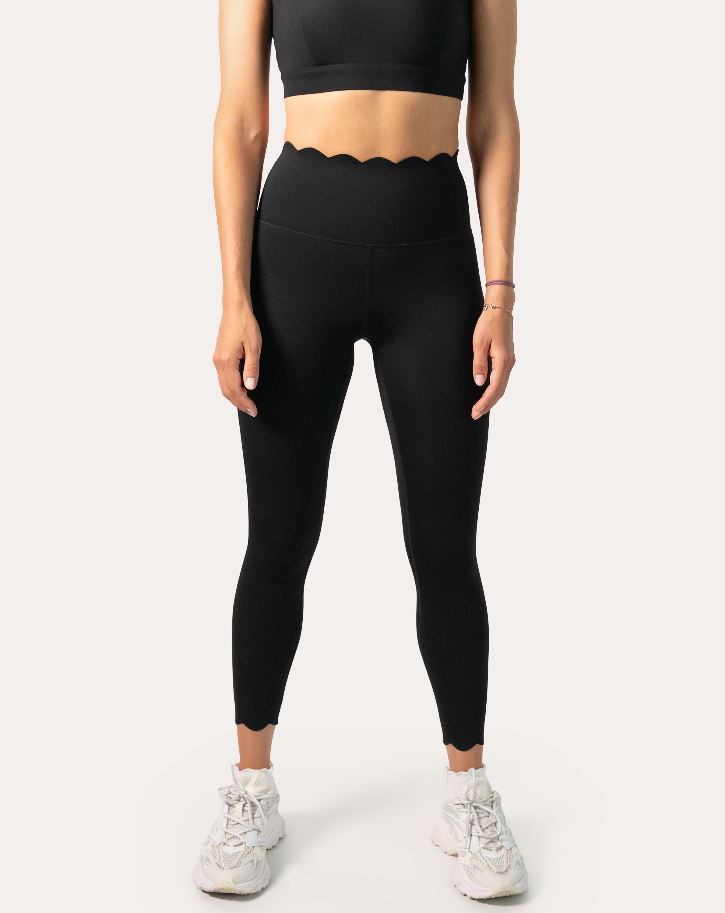 Front view of a woman wearing scallop high-waist black gym leggings, buttery soft second-skin fit with ankle length, styled with white trainers