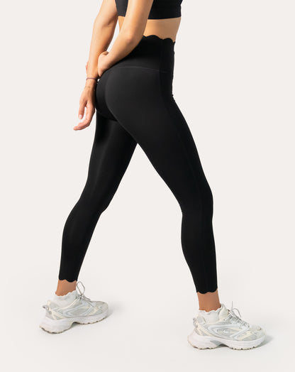 Side lunge pose showing sculpting fit of scallop-waist black leggings with smooth four-way stretch and a neat scalloped hem