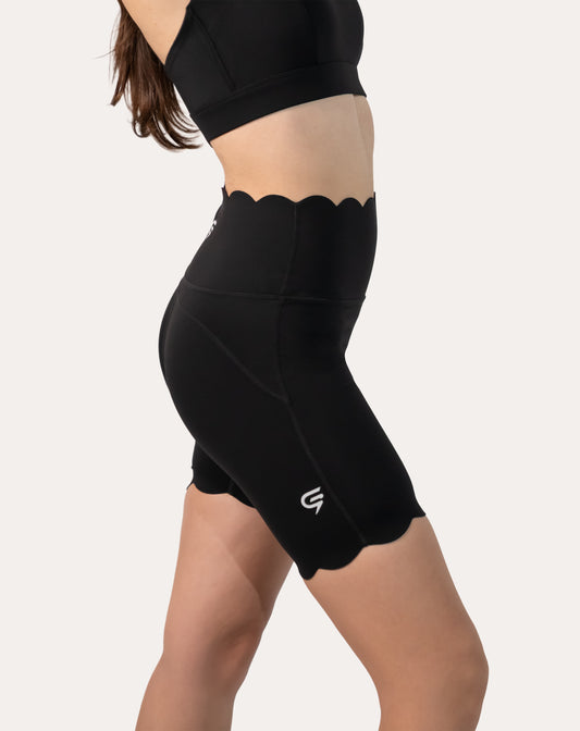 Left three-quarter close-up of black workout shorts with scalloped high waist, scalloped hem, and side pocket seam