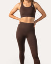 Full-body studio shot of a woman in a brown sports bra and matching leggings, hands on hips