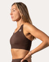 Side profile close-up highlighting wide straps, smooth scoop neck, chest logo, and quick-dry fabric of the brown sports bra