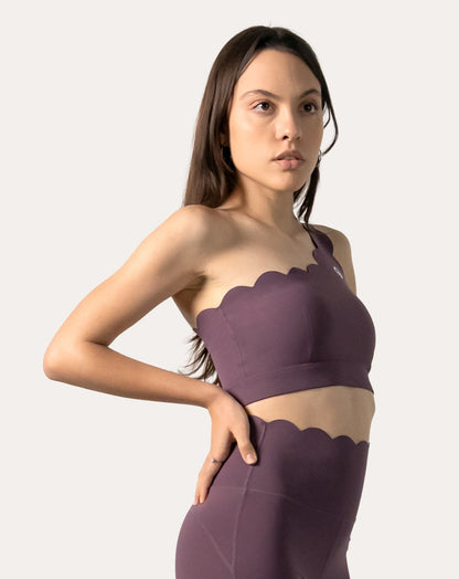 Front three-quarter view, hands on hips, purple sports bra with scalloped one-shoulder neckline