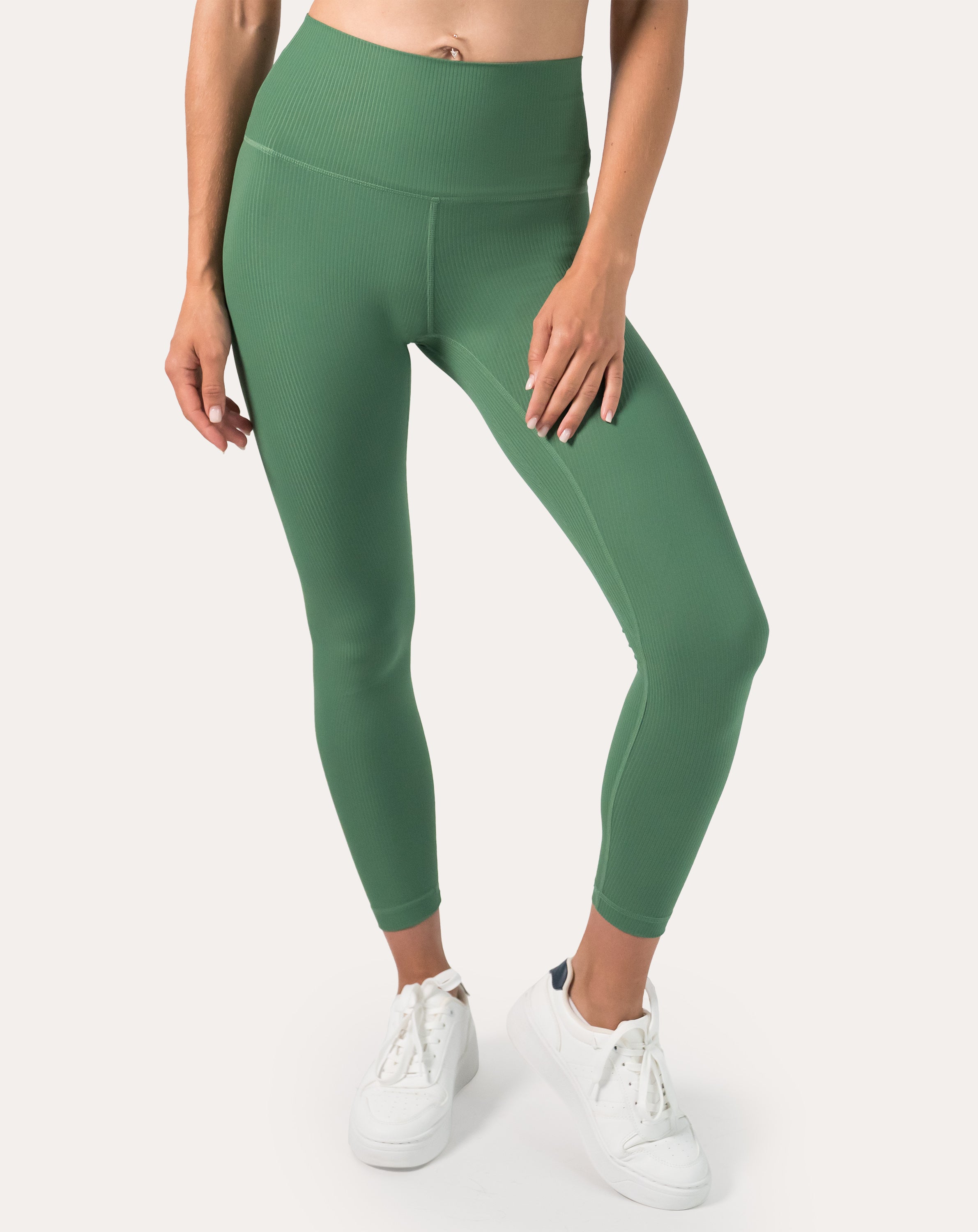 Close front view of women’s ribbed leggings in green with a wide high-waist band; seamless, ankle-length fit styled with white trainers