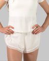 Front view, in motion, of white women’s 2-in-1 running shorts with skort-style side split overlay and drape; model’s hands on hips.
